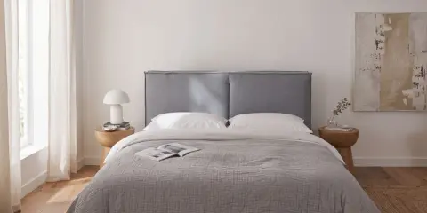 Sypialnia z łóżkiem w kolorze popielatym, białą pościelą i zasłonami. Na stoliku nocnym lampka i książka.