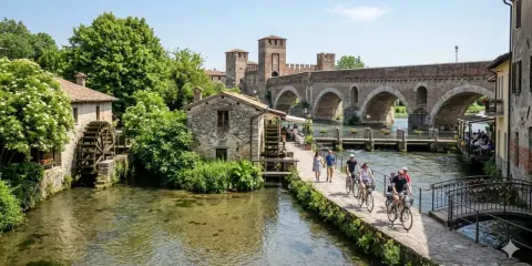 Un pittoresco borghetto sul Mincio con mulini ad acqua, un ponte ad archi e persone che passeggiano e pedalano.