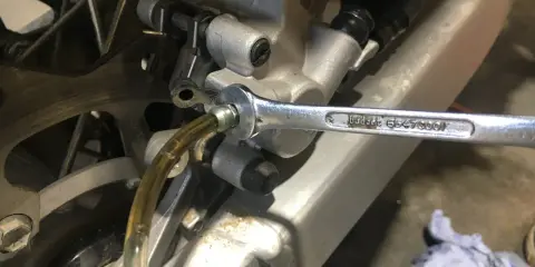 Klucz nasadowy odkręca odpowietrznik zacisku hamulcowego motocykla, podczas gdy przez przezroczysty wężyk płynie płyn. To etap wymiany płynu hamulcowego.