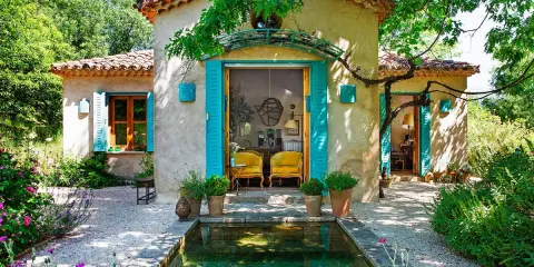 Encantadoras fachadas de casas rústicas con contraventanas turquesas y un estanque rodeado de flores.