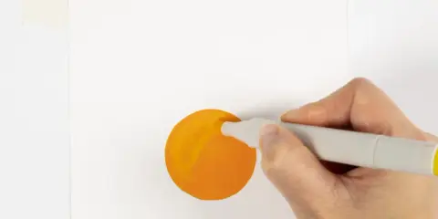 En hand håller en grå tuschpenna och målar en orange cirkel på vitt papper.