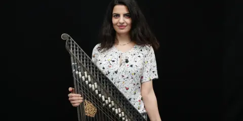 Une musicienne tient un kanoun, instrument de musique à cordes pincées, devant un fond noir.