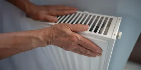 Manos sobre un radiador blanco, sintiendo el calor. El radiador hace ruido, quizás por el agua circulando.