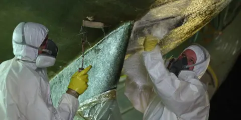 Zwei Personen in Schutzkleidung reparieren den Rumpf eines Bootes. Sie tragen Glasfasermatten auf, um Osmose-Schäden zu beheben.