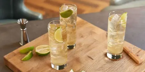 Trzy orzeźwiające whisky ze spritem, udekorowane plasterkami limonki, stoją na drewnianej desce. Obok leżą świeże kawałki limonki.