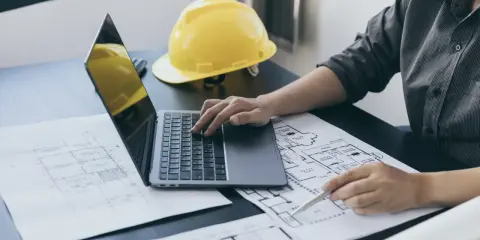 Architekt analizuje plany, porównując powierzchnię zabudowy z użytkową. Na biurku laptop, żółty kask i rysunki techniczne.