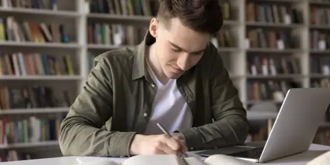 Młody mężczyzna w bibliotece pochyla się nad książkami i laptopem, pisząc notatki. Zastanawia się, czy technikum wlicza się do stażu pracy do emerytury.