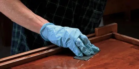 Mano enguantada lijando superficie de madera barnizada. El lijado es un paso clave para aplicar lac.
