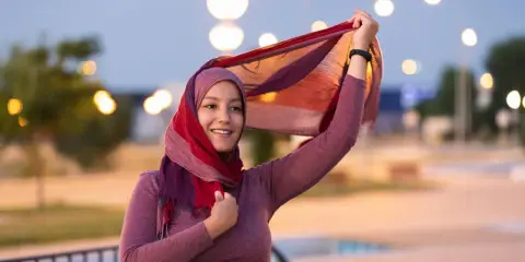 Lächelnde muslimische Frau mit buntem Hijab und Schal, der im Wind weht. Ein Bild, das die Vielfalt und Schönheit islamischer Kultur zeigt.