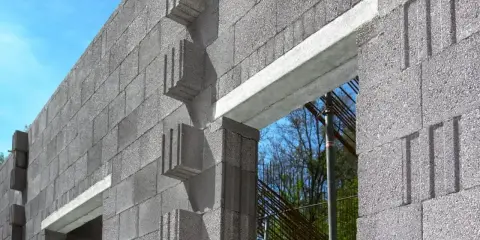 Ściana z bloczków betonowych, widoczny betonowy nadproże nad otworem okiennym. W tle zbrojenie i drzewa.