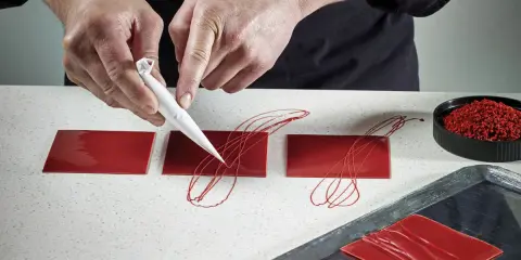 Pâtissier décorant des carrés de chocolat rouge avec un cornet. Le papier guitare, c'est quoi ? C'est ce support qui permet ces décorations fines.