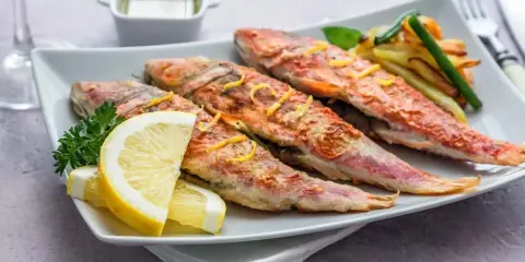 Cuatro salmonetes al horno, dorados y jugosos, servidos con rodajas de limón, perejil y patatas fritas.