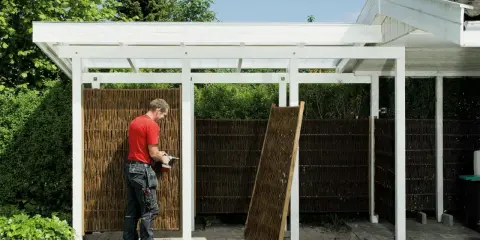 Man bygger redskapsbod med flätade paneler. En ny panel står lutad mot stommen.