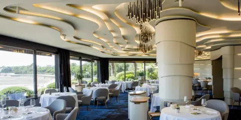 Salle élégante d'un restaurant chef de baie, tables dressées, vue sur la mer et jardin luxuriant. Plafond ondulé et luminaires modernes.