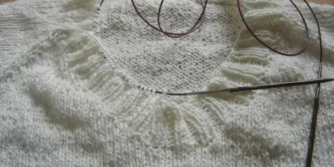 Biały sweter w trakcie dziergania, widoczny fragment kołnierza i druty. Jak zrobić kołnierz do swetra na drutach?