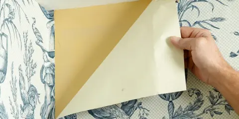 Ręce odklejają tapetę z niebieskim wzorem w narożniku. Pod spodem widać żółtą ścianę.