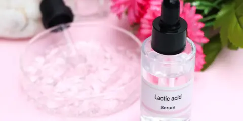 Serum z kwasem mlekowym w szklanej buteleczce z pipetą, obok różowych kwiatów i naczynia laboratoryjnego.