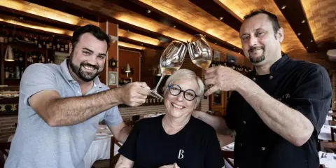 Dos hombres y una mujer brindan con copas de vino en Ca la Pepa, un restaurante acogedor con estanterías de vino.