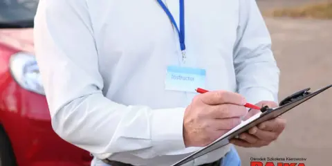 Instruktor notuje dopuszczalne błędy na egzaminie praktycznym na prawo jazdy. Obok czerwony samochód.