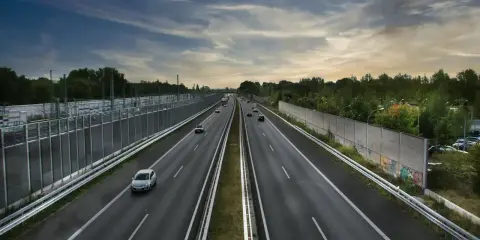 Szeroka autostrada z samochodami, wjazd na autostradę w tle, po bokach ekrany akustyczne i drzewa.