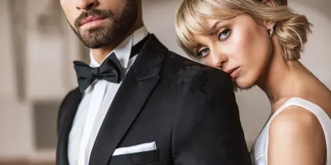 Elegancki mężczyzna w czarnym smokingu z muchą i białą koszulą. Obok niego opiera się kobieta w białej sukni. Idealny przykład, jaka koszula do muchy.