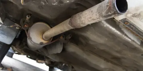 Vista inferior de un coche, mostrando el sistema de escape con un silenciador y tuberías oxidadas. El colector de escape es una parte crucial del sistema.