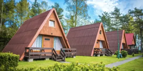 Ustronny domek nad jeziorem, jeden z kilku drewnianych domków w kształcie litery A, otoczony zielenią i drzewami.