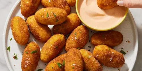 Croquetas de patata doradas y crujientes, espolvoreadas con perejil y sal, listas para ser mojadas en una cremosa salsa rosa.