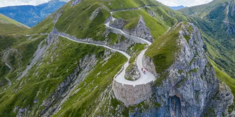 Un tortuoso cammino del sale si snoda tra montagne verdi, con ciclisti che affrontano tornanti mozzafiato su un sentiero panoramico.