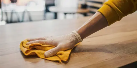 Ręka w rękawiczce wykonuje ścieranie kurzu z drewnianego blatu za pomocą żółtej ściereczki.