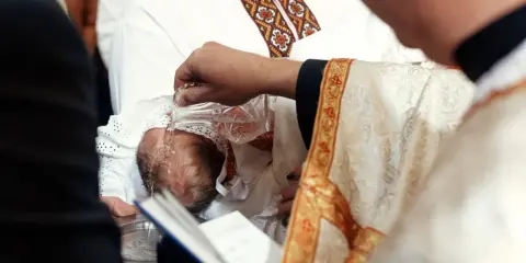 Chrzestni słuchają słów kapłana, gdy ten polewa wodą głowę niemowlęcia.
