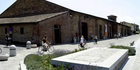 Passeggiata in bicicletta davanti ai Magazzini del Sale di Cervia, con persone che pedalano su un lungomare acciottolato, tra aiuole fiorite e antichi edifici in mattoni.