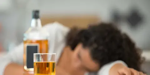 Szklanka z alkoholem i butelka na stole, osoba śpiąca w tle.