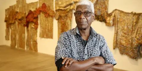 A distinguished man with white hair and glasses, arms crossed, stands before a wall adorned with shimmering, textured artworks, a testament to the enduring legacy of famous artists today.