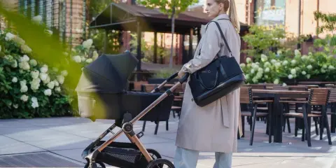 Elegancki wózek dla chłopca, prowadzony przez kobietę w beżowym płaszczu, spaceruje po urokliwym zaułku z kwitnącymi hortensjami.