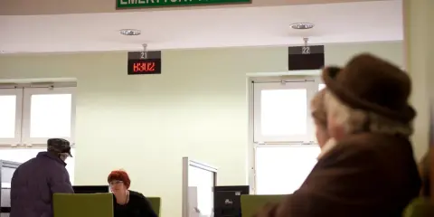 Ludzie czekają na obsługę w urzędzie "Emerytury i Renty". Zastanawiają się, w jakim miesiącu najlepiej przejść na emeryturę.