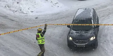 En räddningsarbetare spärrar av ett område med band. Snö faller och täcker marken och en bil. Kanske har något ruttnande material hittats.