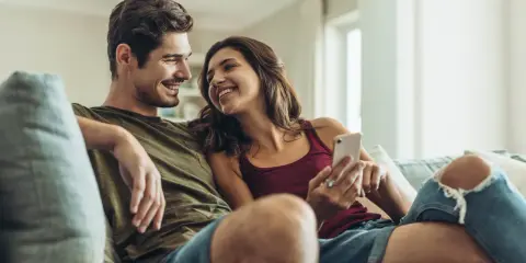 Pareja sonriente en el sofá, compartiendo un momento íntimo. Ella le muestra algo en su móvil, una forma de como expresar sentimientos.
