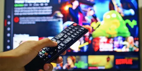 Dłoń trzymająca pilot do telewizora, na ekranie menu platformy streamingowej. Wady i zalety reklamy telewizyjnej – czy warto ją oglądać?