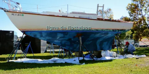 Un bateau est sur des chandelles, prêt pour une peinture. La ligne de flottaison est visible sur la coque bleue. Un homme travaille sur le bateau.