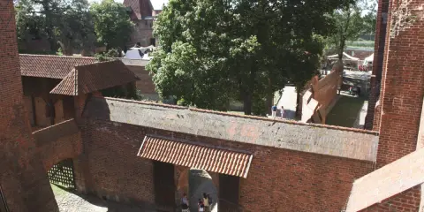 Wielki zamek w Malborku, ceglane mury, dziedziniec z drzewami i ludźmi spacerującymi pośród zabytkowej architektury.