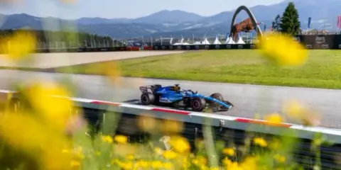Un coche de F1 azul compite en un circuito soleado, con flores amarillas en primer plano. ¡Un gran resultado f1 hoy!