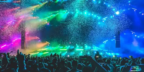 Folla in delirio a un concerto a Rimini, con coriandoli colorati e luci psichedeliche che illuminano la scena.