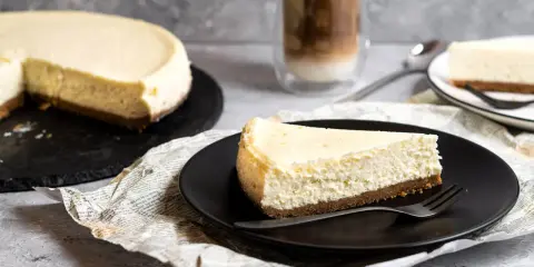 Ein Stück New York Cheesecake auf schwarzer Platte, daneben ein ganzer Kuchen und ein Latte Macchiato.