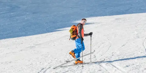 Uśmiechnięty mężczyzna w pomarańczowej kamizelce i niebieskich spodniach na nartach, gotowy na skitury dla początkujących.