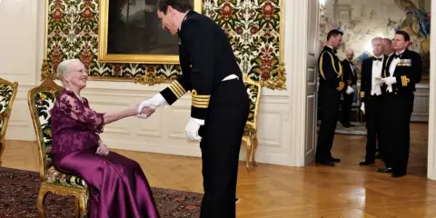 Drottning Margrethe, trots att hon är sjuk, tar emot en officer med ett leende. Hon sitter i en elegant stol, klädd i en lila klänning.