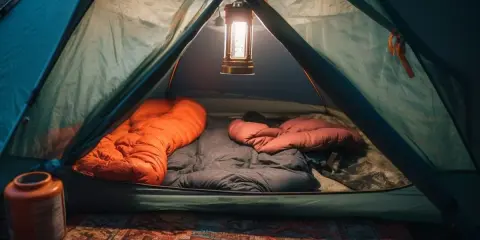 Wnętrze namiotu gotowe do spania. Dwa śpiwory, pomarańczowy i brązowy, czekają na noc. Lampion rzuca ciepłe światło.