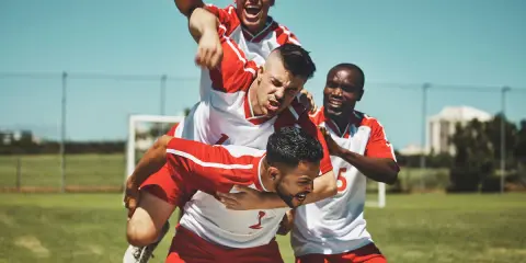 Piłkarze świętują zwycięstwo. Jeden z nich niesie kolegę na barana, krzycząc z radości. To może być celebracja hattricka, czyli zdobycia trzech bramek w jednym meczu.