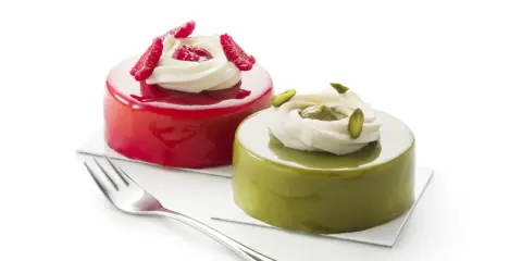 Deux petits gâteaux brillants, l'un rouge vif, l'autre vert avec un glaçage pistache, ornés de crème et de fruits.