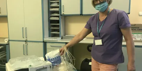 Pielęgniarka w maseczce pokazuje zestaw do cado dializa. Na stole aparat i materiały medyczne.